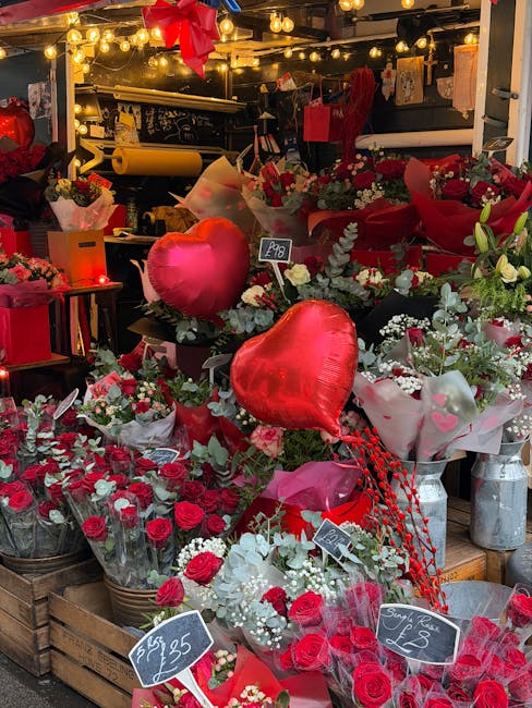 An array of fresh floral arrangements and bouquets displayed at a flower shop, featuring red roses, white lilies, and mixed greenery. The display includes heart-shaped foil balloons, wrapped in clear 