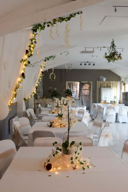 An elegantly decorated indoor venue with long banquet tables draped in white tablecloths, adorned with small floral arrangements featuring white orchids and greenery in glass vases. The tables are sur