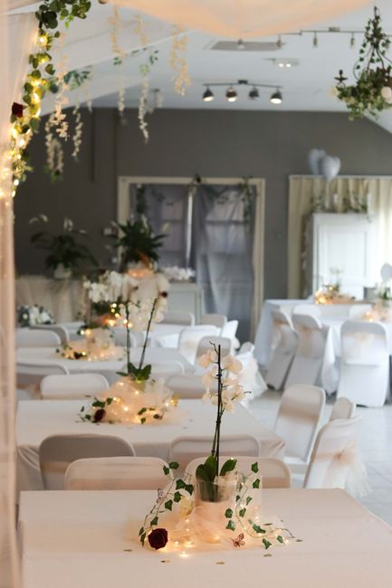 A floral arrangement featuring a tall, slender vase holding a delicate bouquet of white orchids and greenery, set on a white tablecloth. Surrounding the vase are string lights and small white flowers,