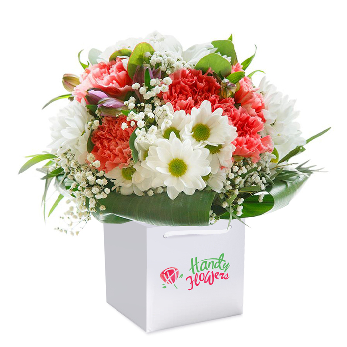 Displayed upright within a sleek, square white gift bag, this vibrant floral arrangement crafted for Hatch End captures both the energy of local high roads and the serenity found in nearby parks. At its heart, layers of coral-pink and radiant red carnations show off their signature ruffled petals, adding plush volume and softness that seem almost tangible in the gentle light. Bright white daisy-like blooms sparkle throughout, each with a central burst of yellow-green, acting as luminous highlights and balancing the composition's warmth. Gentle sprigs of pure white gypsophila-baby's breath-float around the primary flowers, enhancing the bouquet with lightness and subtle intricacy. A handful of closed, purple-tipped buds nestle on the left, promising newness and dimension. Surrounding all are abundant, glossy green leaves, their deep hues forming a sturdy, elegant support and echoing the lush greenery of Hatch End's suburban streets. The arrangement is thoughtfully rounded, its fullness suggesting generosity and attentive design. The white hand-tied bag, with sturdy ribbon handles, speaks to a practical yet stylish approach-a nod to the convenience of local delivery. This composition beautifully represents the vibrant spirit and floral artistry of Hatch End, offering a memorable gift for birthdays, anniversaries, or personal milestones, all lovingly assembled for the community.