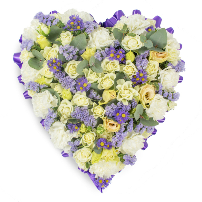 A meticulously arranged heart of sympathy flowers rests against a pristine white surface, its compact face telling a story of comfort in muted cream, pale yellow and lavender tones. The eye is drawn first to the many cream and pale yellow roses packed closely together - some in tight button form, others unfurling into soft, luxurious cups with a slight hint of green at their throat - which create a plush, tactile center. Surrounding and lifting the roses are generous sprays of white carnations, their layered ruffles adding mass and a traditional note of condolence. Throughout the piece, small clusters of lavender-hued statice and vivid purple, aster-like blooms with clear yellow centers add cool, contrasting punctuation points that make the composition feel both delicate and complete. The heart's outline is neatly finished with a continuous band of deep purple satin ribbon, folded into a refined border that reinforces the shape and gives the arrangement an understated elegance. Intermingled among the florals are round, blue-green eucalyptus leaves whose soft sheen and distinctive silhouettes ease the density, introduce a calming green note and imply a fresh, subtle scent. Bathed in gentle, neutral lighting, the tribute reads as peaceful and reverent - an appropriate floral expression for memorial services, chapel placements, or graveside dedications in Hatch End, near Harrow and the surrounding communities - offering a quiet, heartfelt symbol of remembrance and enduring affection.