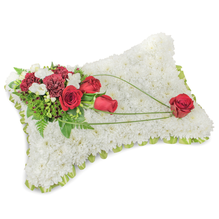 A sorrowful yet serenely beautiful rectangular cushion of blossoms, this traditional funeral pillow is created from hundreds of pristine white chrysanthemums packed densely to form a velvety, cloud-like surface. Along the softly rounded edges a pale green ribbon or slender foliage outline is visible, giving the cushion a gentle, framed silhouette that feels both simple and carefully attended. The upper left corner is embellished with a lush spray of deep red roses and rich burgundy carnations; the roses range from tight, hopeful buds to fully opened blooms whose velvety layers convey warmth and reverence. Scattered among them are small, pale white accent blooms-delicate freesias or tiny spray chrysanthemums-that provide luminous contrast and a light, fragrant whisper. Supporting foliage includes feathered fern sprigs and broader green leaves that deepen the composition and make the reds read even more vivid. Across the white chrysanthemum plane, three arcs of bright green bear-grass or similarly supple foliage create a graceful diagonal movement, leading the gaze to a single, immaculate red rose positioned on the lower right corner of the pillow, as if gently laid there by a loved one. The light in the photograph is soft and even, throwing minimal shadow and emphasizing petal texture and tone; the aroma is subtle, a respectful, clean floral scent rather than a perfume. This arrangement carries the traditional language of condolence-peace, purity and enduring love-and is the kind of memorial crafted by local florists for services at community churches, chapels or gravesides in and around Hatch End and Pinner.